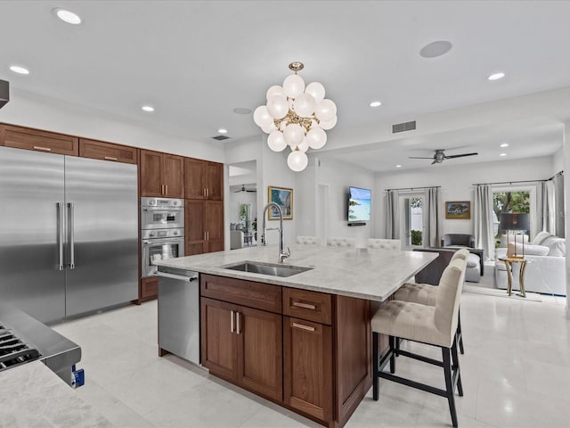 kitchen with ceiling fan with notable chandelier, a kitchen breakfast bar, a center island with sink, sink, and stainless steel appliances