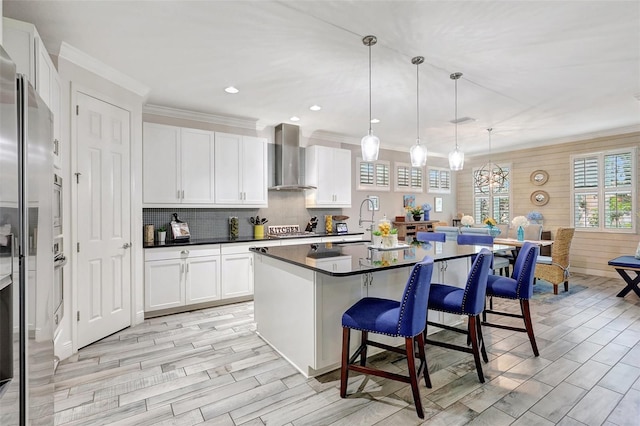 kitchen featuring white cabinetry, wall chimney range hood, an island with sink, and decorative light fixtures