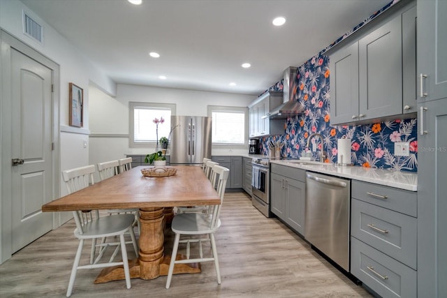 kitchen with appliances with stainless steel finishes, wall chimney exhaust hood, gray cabinets, light hardwood / wood-style flooring, and sink