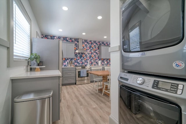 kitchen with appliances with stainless steel finishes, gray cabinetry, wall chimney exhaust hood, stacked washer / drying machine, and light hardwood / wood-style flooring