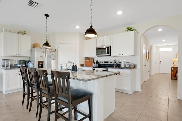 kitchen with light stone countertops, stainless steel appliances, a center island with sink, white cabinets, and hanging light fixtures