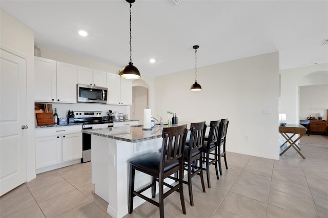 kitchen featuring a kitchen bar, appliances with stainless steel finishes, dark stone counters, a kitchen island with sink, and decorative light fixtures