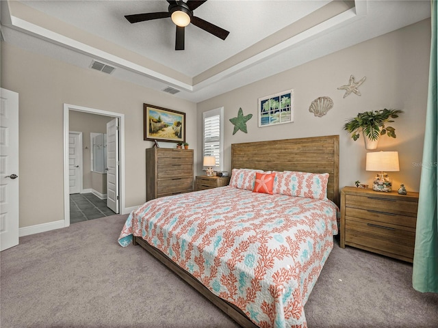 bedroom with dark colored carpet, ensuite bathroom, a tray ceiling, and ceiling fan