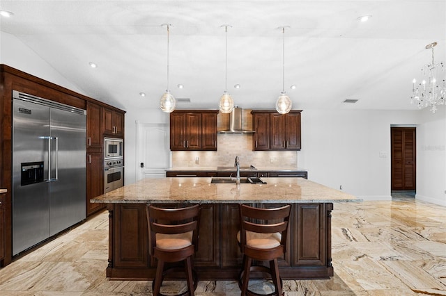 kitchen featuring light stone countertops, an island with sink, hanging light fixtures, wall chimney exhaust hood, and built in appliances