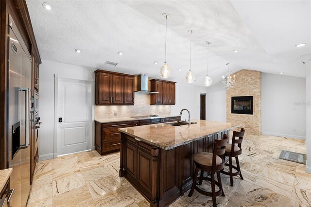 kitchen featuring hanging light fixtures, an island with sink, vaulted ceiling, wall chimney exhaust hood, and light stone counters