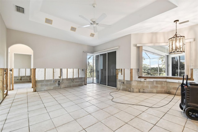 tiled empty room with ceiling fan with notable chandelier, a tray ceiling, and tile walls