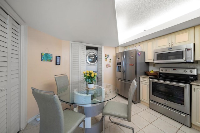 kitchen with stacked washer and clothes dryer, cream cabinetry, stainless steel appliances, light tile patterned floors, and a textured ceiling