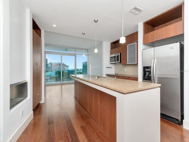 kitchen with a kitchen island with sink, sink, light wood-type flooring, appliances with stainless steel finishes, and decorative light fixtures