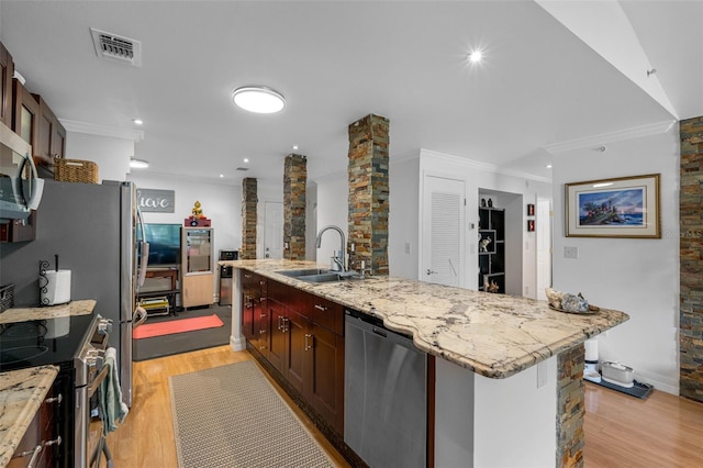 kitchen with appliances with stainless steel finishes, sink, light wood-type flooring, ornate columns, and a kitchen island with sink