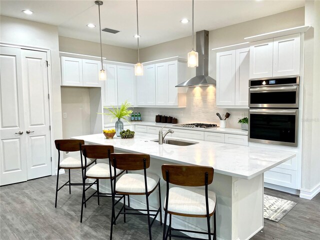 kitchen featuring an island with sink, appliances with stainless steel finishes, decorative light fixtures, wall chimney exhaust hood, and sink