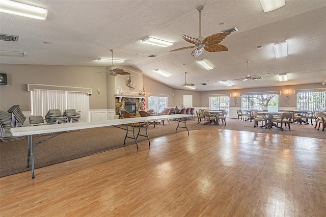 game room featuring a stone fireplace, a textured ceiling, ceiling fan, vaulted ceiling, and light hardwood / wood-style flooring