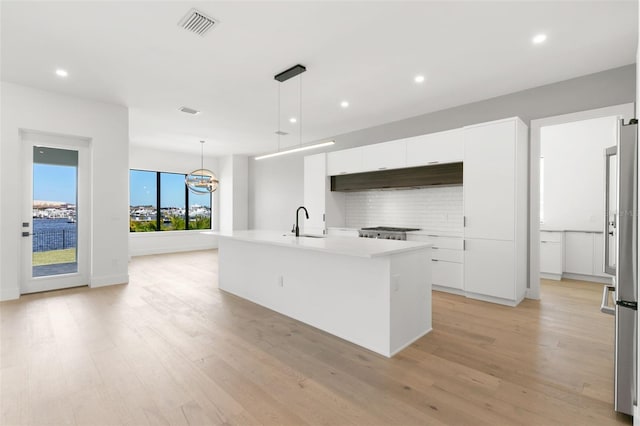 kitchen with decorative backsplash, white cabinets, hanging light fixtures, a center island with sink, and light hardwood / wood-style flooring