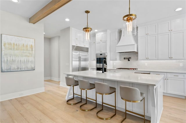 kitchen with appliances with stainless steel finishes, white cabinetry, a kitchen island with sink, light wood-type flooring, and decorative light fixtures