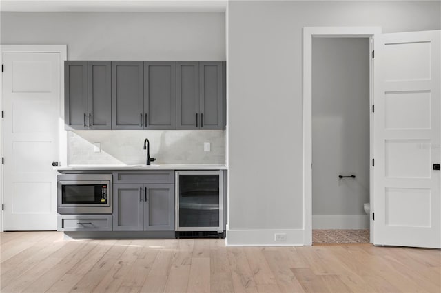 bar featuring tasteful backsplash, beverage cooler, stainless steel microwave, light hardwood / wood-style flooring, and gray cabinets