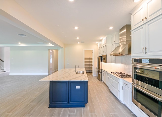 kitchen with wall chimney range hood, a center island with sink, sink, light stone countertops, and white cabinetry