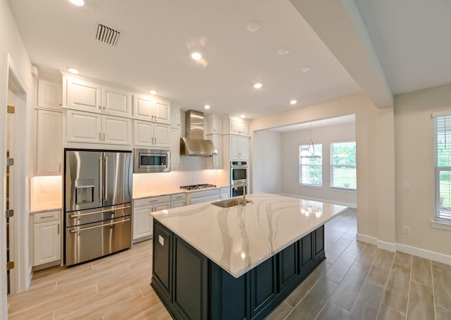 kitchen with wall chimney range hood, white cabinets, an island with sink, sink, and stainless steel appliances