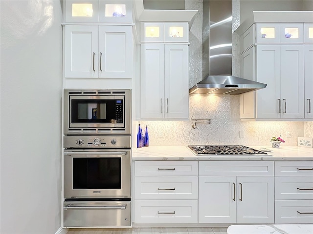 kitchen with wall chimney range hood, tasteful backsplash, light stone counters, white cabinetry, and stainless steel appliances