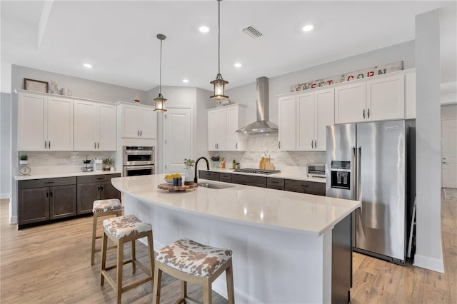 kitchen featuring wall chimney exhaust hood, stainless steel appliances, an island with sink, and white cabinets