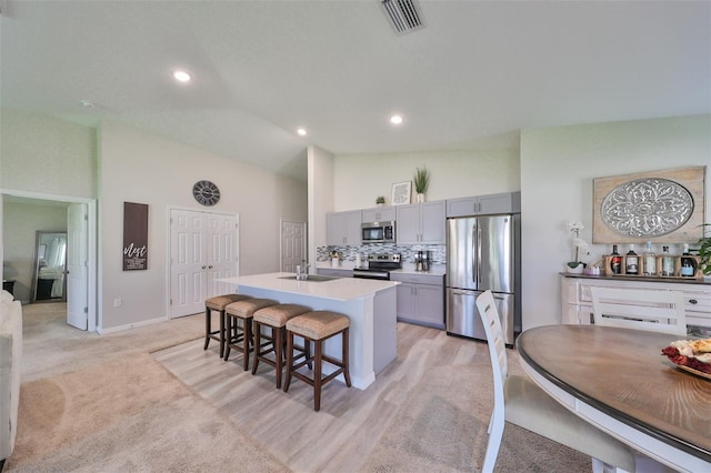 kitchen featuring a kitchen island with sink, light carpet, a kitchen bar, gray cabinets, and appliances with stainless steel finishes