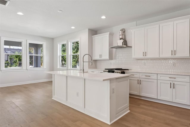 kitchen with white cabinetry, wall chimney range hood, light hardwood / wood-style floors, and an island with sink