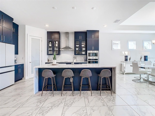 kitchen with a breakfast bar, an island with sink, wall chimney range hood, and white fridge