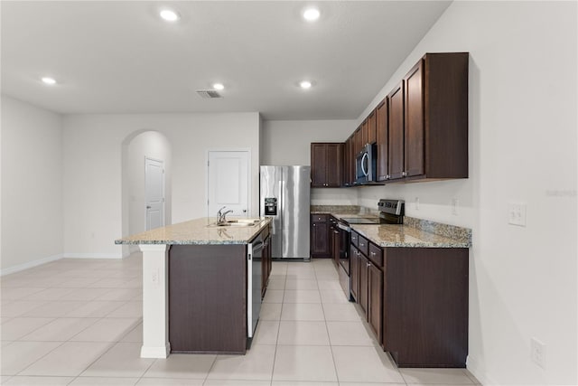 kitchen with light stone countertops, sink, dark brown cabinets, stainless steel appliances, and a kitchen island with sink