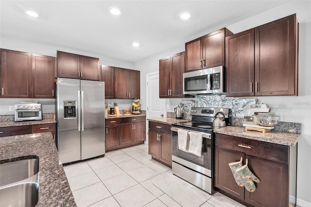 kitchen with appliances with stainless steel finishes, sink, dark brown cabinets, dark stone counters, and light tile patterned floors