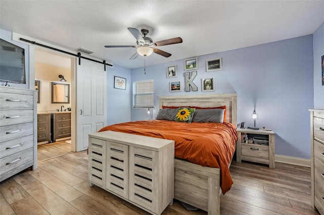 bedroom with ensuite bath, a barn door, light hardwood / wood-style floors, and ceiling fan