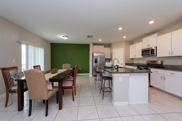 kitchen featuring dark stone counters, stainless steel appliances, a kitchen island with sink, light tile patterned floors, and white cabinetry