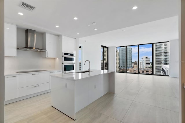 kitchen featuring a kitchen island with sink, sink, wall chimney exhaust hood, double oven, and white cabinetry