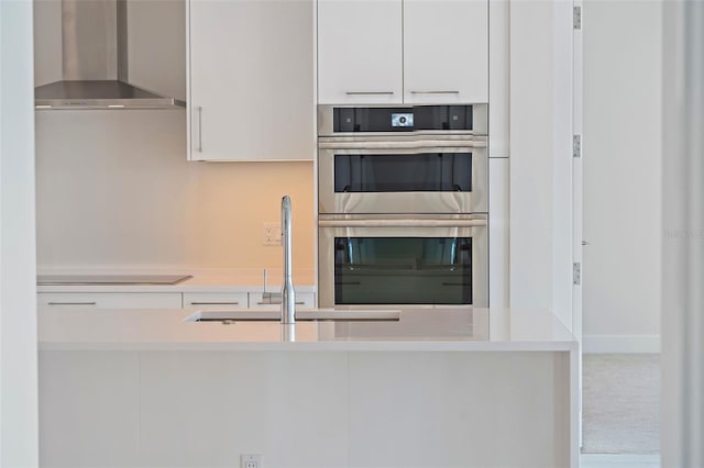 kitchen with stainless steel double oven, sink, wall chimney range hood, stovetop, and white cabinetry