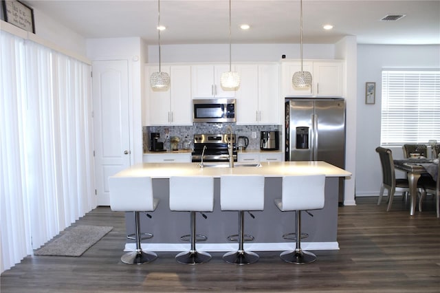 kitchen featuring dark wood-type flooring, an island with sink, white cabinets, decorative light fixtures, and appliances with stainless steel finishes