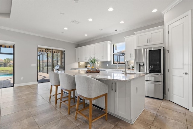 kitchen with backsplash, refrigerator with glass door, white cabinets, a kitchen island with sink, and light stone countertops