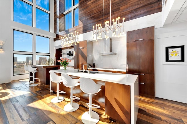 kitchen featuring wall chimney range hood, a kitchen breakfast bar, a notable chandelier, a towering ceiling, and an island with sink