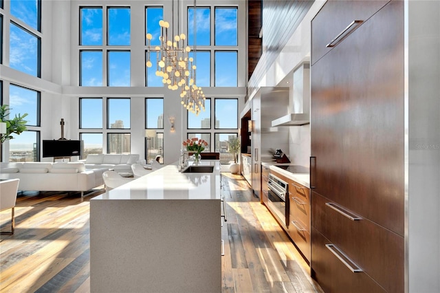 kitchen featuring stainless steel oven, wall chimney exhaust hood, a high ceiling, a notable chandelier, and a kitchen island with sink