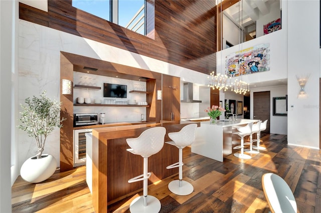 bar featuring beverage cooler, wood-type flooring, wall chimney range hood, and a towering ceiling