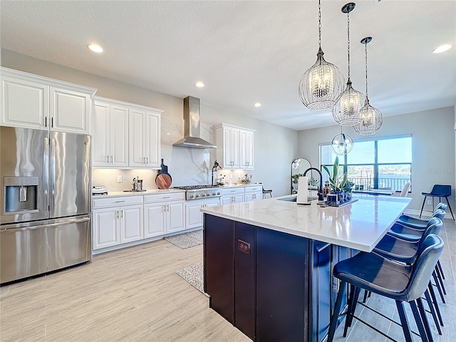 kitchen featuring wall chimney range hood, appliances with stainless steel finishes, white cabinetry, a kitchen island with sink, and decorative light fixtures