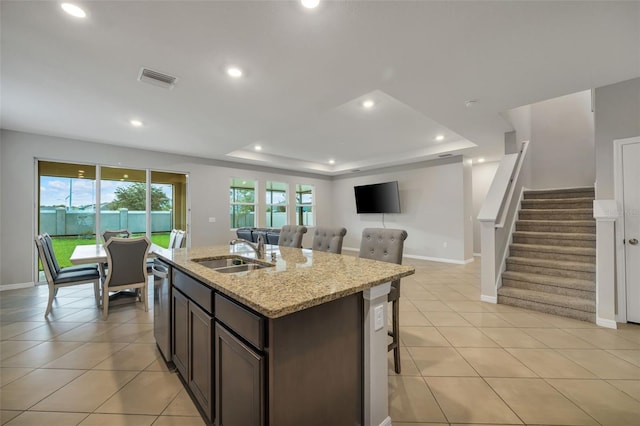 kitchen with a raised ceiling, sink, stainless steel dishwasher, light stone countertops, and an island with sink