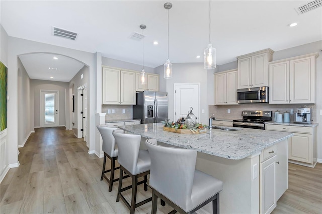 kitchen featuring hanging light fixtures, stainless steel appliances, a center island with sink, light stone countertops, and light hardwood / wood-style floors