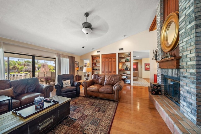 living room with ceiling fan, a textured ceiling, a brick fireplace, light hardwood / wood-style flooring, and lofted ceiling with beams