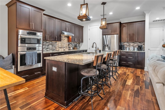 kitchen with sink, appliances with stainless steel finishes, a kitchen island with sink, hanging light fixtures, and light stone countertops