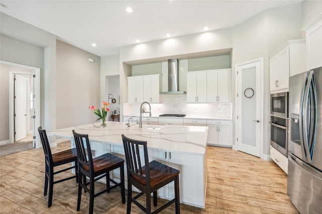 kitchen featuring white cabinets, an island with sink, stainless steel appliances, wall chimney exhaust hood, and sink