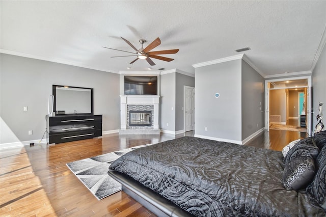 bedroom with a stone fireplace, hardwood / wood-style floors, crown molding, a textured ceiling, and ceiling fan