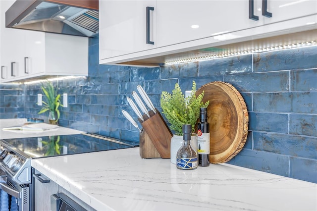 room details featuring white cabinets, tasteful backsplash, stainless steel range oven, wall chimney exhaust hood, and light stone counters