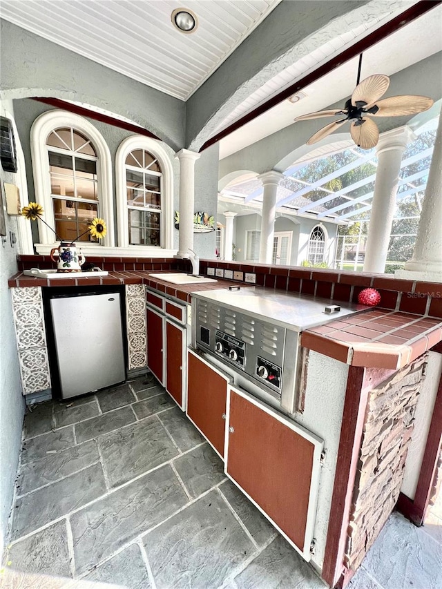 kitchen with tile countertops, sink, ceiling fan, fridge, and decorative columns