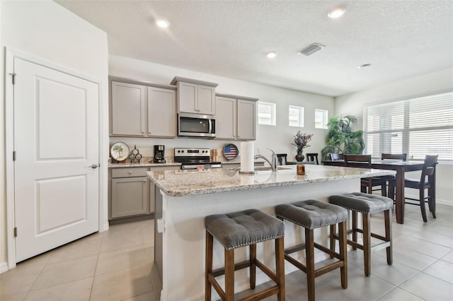 kitchen with light tile patterned floors, appliances with stainless steel finishes, a kitchen island with sink, gray cabinetry, and light stone countertops