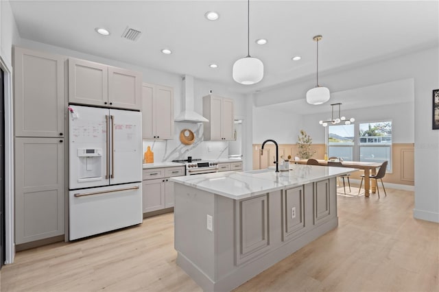 kitchen featuring white fridge with ice dispenser, a kitchen island with sink, sink, wall chimney exhaust hood, and decorative light fixtures