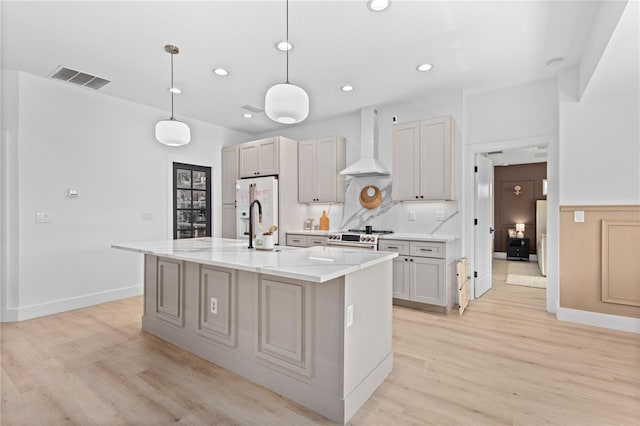 kitchen with light wood-type flooring, wall chimney range hood, an island with sink, and hanging light fixtures