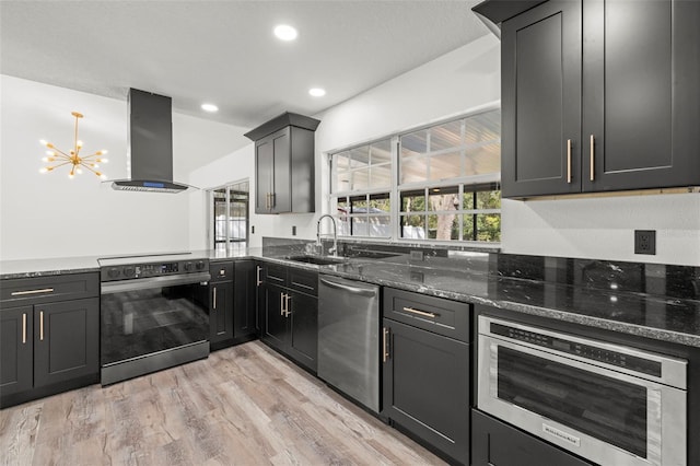 kitchen featuring wall chimney range hood, appliances with stainless steel finishes, dark stone countertops, light hardwood / wood-style floors, and sink