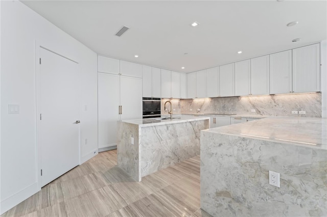 kitchen with sink, tasteful backsplash, light stone countertops, an island with sink, and white cabinets